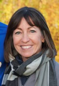 Photograph of a smiling white woman looking at the camera with a yellow background, shoulder-length brown hair, wearing a grey scarf