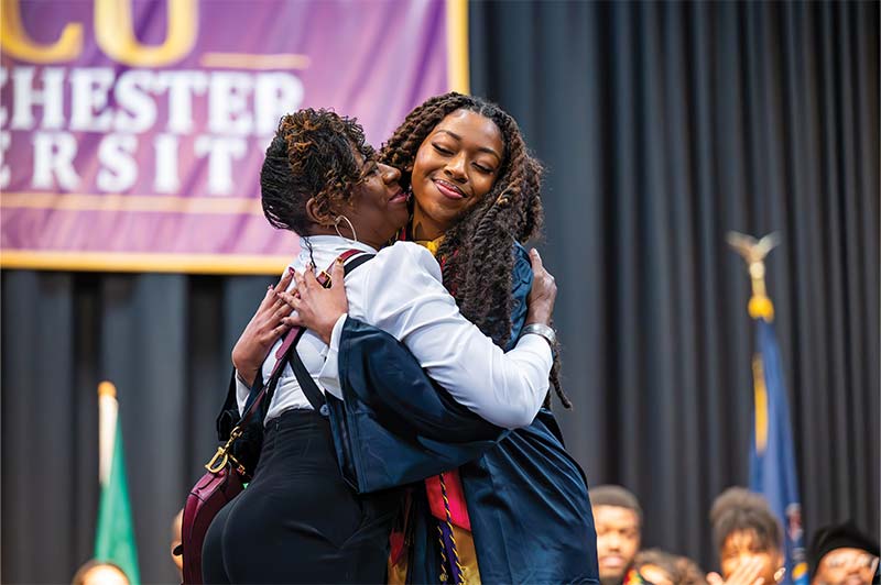 A graduate in cap and gown hugs another person on stage during a university ceremony, both smiling warmly.