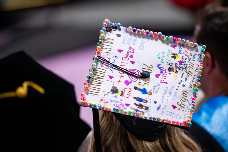 Close-up of a decorated graduation cap covered in colorful beads, drawings, and handwritten messages. The year “2025” is written on top, and people are seated in the background.
