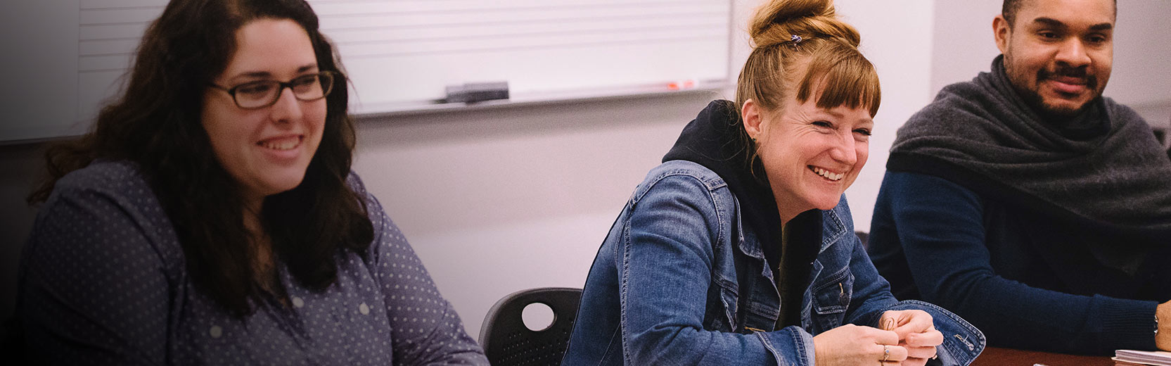 Three adult students sit together in a classroom, smiling and engaged in discussion, with a whiteboard in the background.
