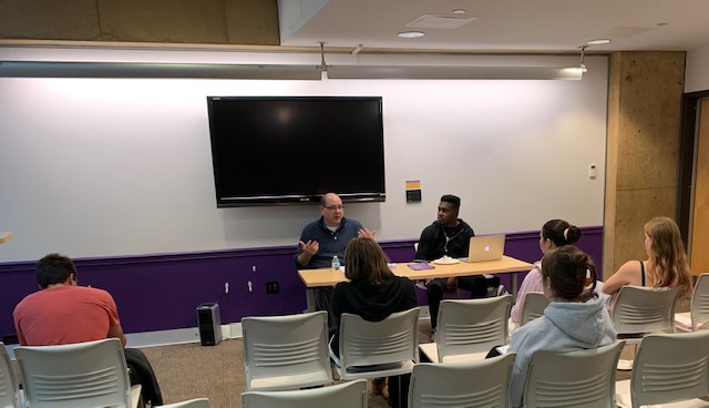 Students sitting in a classroom. Two presenters at the front sitting at a table.