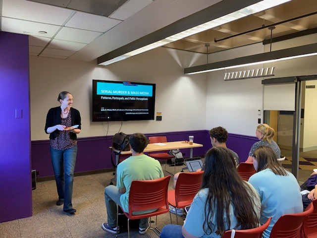 Woman walking down the perimeter of the room. Students sitting looking at a screen at the front.